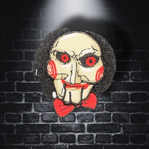 May include: Embroidered patch featuring a stylized face with red eyes, a red bow tie, and black hair. The patch has a cream-colored face and is set against a dark brick wall background. The design is reminiscent of a horror movie character.