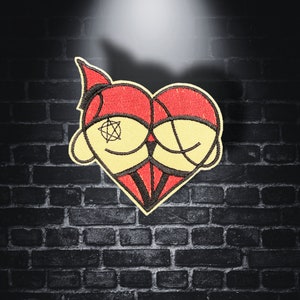 Pode incluir: Um coração bordado em vermelho e amarelo com um pentagrama preto dentro de um olho e um contorno preto. O coração é costurado com fio preto e tem um padrão de listras vermelhas e pretas.