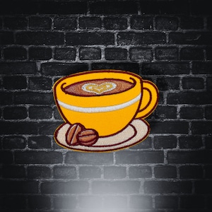 Può includere: Toppa ricamata raffigurante una tazza di caffè gialla con una striscia bianca e un disegno a forma di cuore nel caffè. Due chicchi di caffè sono sul piattino. La toppa è contro un muro di mattoni scuri.