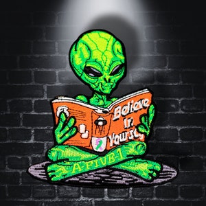 Könnte beinhalten: Grüner Außerirdischer, der im Schneidersitz sitzt und ein Buch liest, auf dem "Glaube an dich selbst" steht. Der Außerirdische trägt ein lila Band am Arm und das Buch ist orange mit schwarzem Text. Der Außerirdische sitzt auf einer grauen Oberfläche mit der Aufschrift "Active 1".