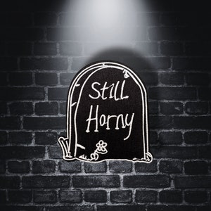 Könnte beinhalten: Ein schwarzer Grabstein mit weißem Text, der "Still Horny" lautet. Der Grabstein steht vor einer dunklen Backsteinmauer.