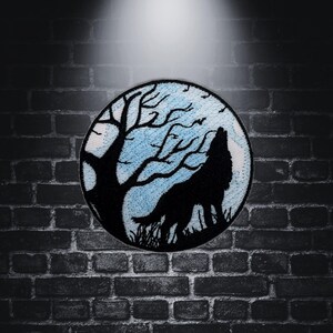 Op de afbeelding: Een zwarte geborduurde patch met een wolf die huilt naar de maan met een boom op de achtergrond. De maan is blauw en wit.