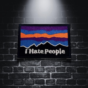 Puede incluir: Parche bordado con borde negro, con un diseño de cordillera con rayas horizontales moradas, naranjas y azules. El texto "I Hate People" está en blanco, debajo del gráfico de la montaña.