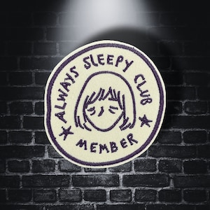May include: Embroidered patch with the text "ALWAYS SLEEPY CLUB MEMBER" in purple on a cream background. The patch features a cartoon illustration of a person with closed eyes. Two stars are on either side of the illustration.