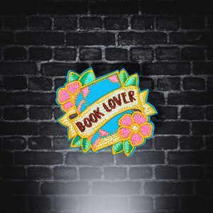 Op de afbeelding: Geborduurde patch met de tekst "BOOK LOVER" op een banner, omringd door roze bloemen en groene bladeren. De patch heeft een lichtblauwe achtergrond en een gele rand. De patch is geplaatst tegen een zwarte bakstenen muur.