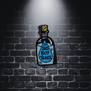 Op de afbeelding: Een blauwe glazen fles met een kurkstop en een blauwe traan met de tekst "Sad Boy Tears" erin. De fles is zwart omlijnd en heeft een witte achtergrond.