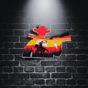 Könnte beinhalten: Gestickter Aufnäher mit einem Cowboy, der auf einem Pferd mit Lasso reitet, vor einem Sonnenuntergangs-Hintergrund. Der Aufnäher ist schwarz umrandet und hat ein rotes, gelbes und oranges Farbschema.