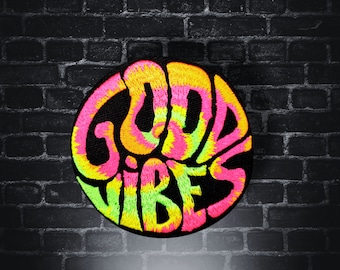 May include: A round, black patch with the words "Good Vibes" in a tie-dye pattern of pink, yellow, and green. The patch is on a black brick wall background.