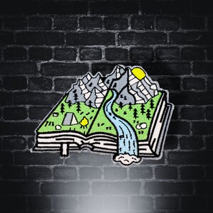 May include: An embroidered patch depicting a book with a mountain range, a river, and a campsite on the pages. The book is open and the pages are curled up at the edges. The mountains are grey and the river is blue. The campsite has a tent and a campfire.