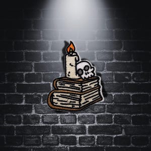 Può includere: Toppa ricamata raffigurante una candela con fiamma, un teschio e due libri impilati. La toppa ha uno sfondo color crema con accenti marroni e arancioni. Il design ha un tema gotico e spettrale.