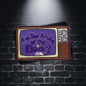 Può includere: Toppa ricamata viola di un televisore vintage con il testo "Are You Afraid of the Dark?" in un contorno bianco.