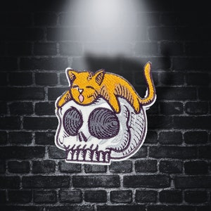 Peut inclure: Écusson brodé représentant un chat de dessin animé reposant sur un crâne. Le chat est orange avec une langue rose, et le crâne est blanc avec des ombres grises. L'écusson a une bordure blanche et est posé sur un mur de briques sombres.