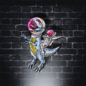 May include: Embroidered patch featuring a blue dinosaur wearing a space helmet, with an astronaut riding on its back. The dinosaur has yellow accents and a pink mouth. The astronaut is in a white spacesuit with a pink helmet. The patch is against a dark brick background.