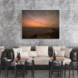 May include: A framed photograph of a night sky with stars and a pink and orange aurora borealis over a body of water. The water is calm and reflects the sky. The photo is hanging on a concrete wall above a brown leather sofa with white cushions.