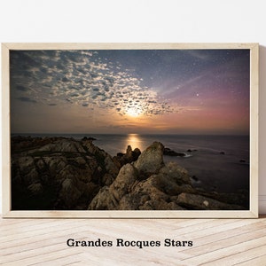 May include: A photograph of a rocky coastline with a full moon rising over the ocean. The sky is filled with stars and clouds. The image is framed in a light wood frame.