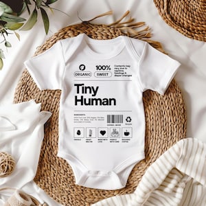 May include: White baby onesie with black text that reads "100% Organic Sweet Tiny Human". The onesie has a label with a list of ingredients, including "Pure Love", "Giggles", "Baby Smiles", "Warm Hugs", "Midnight Cuddles", and "A Dash of Curiosity".