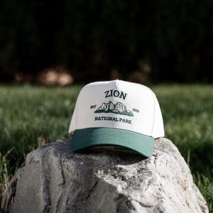 May include: A white and green baseball cap with the text "ZION NATIONAL PARK" and a mountain graphic. The cap has a green brim and is resting on a gray rock. The text includes the year "1919".