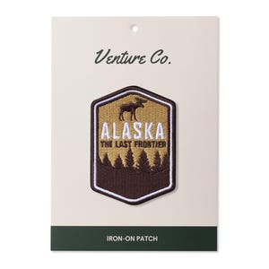 Puede incluir: Parche para planchar con la silueta de un alce marrón sobre un fondo beige con el texto "Alaska The Last Frontier" en blanco. El parche está rodeado por un borde marrón y tiene un contorno blanco. El texto "Venture Co." está impreso en la parte superior de la tarjeta y "Iron-on Patch" está impreso en la parte inferior.