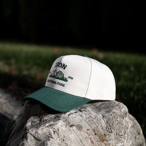 May include: A white and green baseball cap with the text "ZION NATIONAL PARK 1919". The cap has a green brim and a white crown. The cap is sitting on a rock.