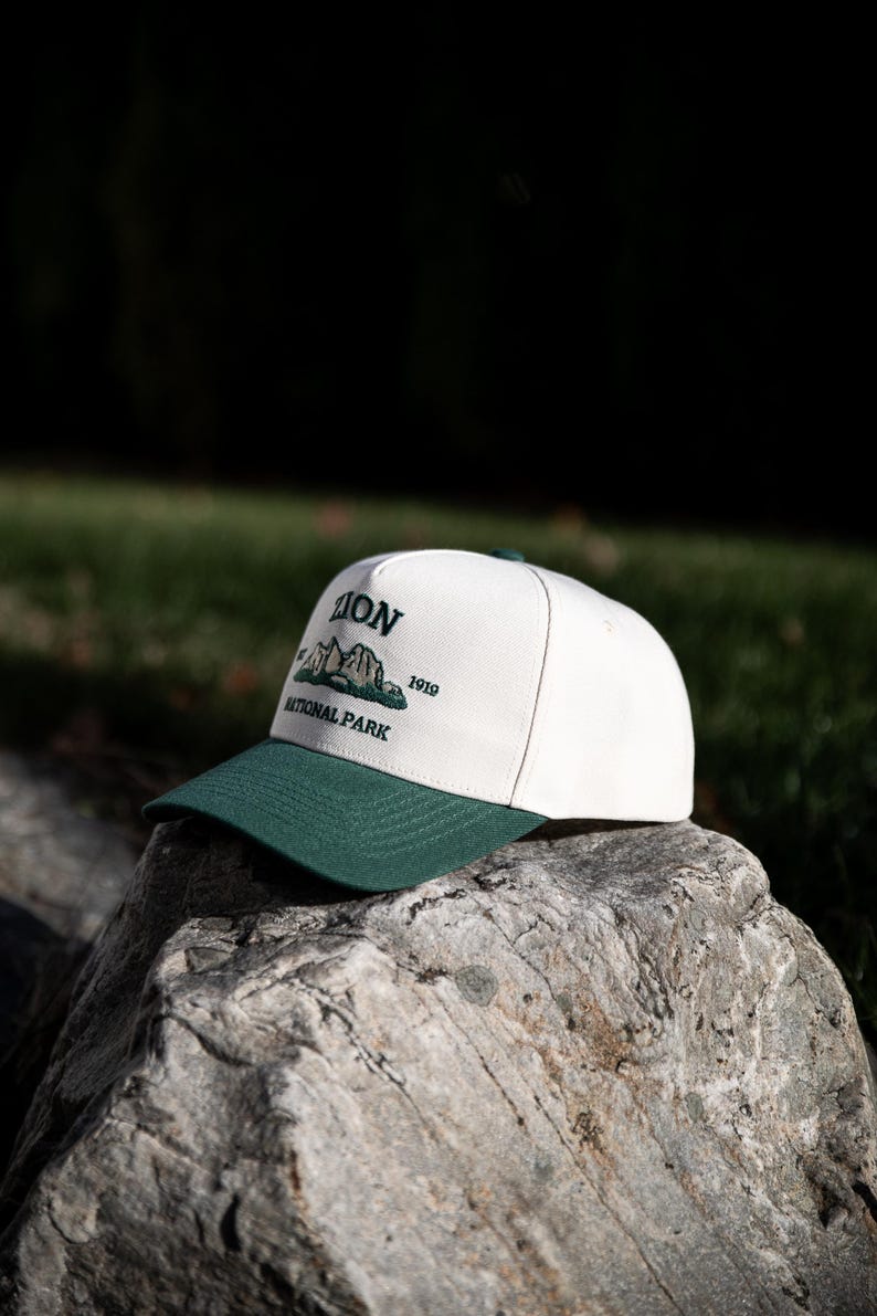 May include: A white and green baseball cap with the text "ZION NATIONAL PARK 1919". The cap has a green brim and a white crown. The cap is sitting on a rock.