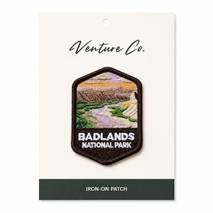 May include: An embroidered patch depicting a landscape from Badlands National Park. The brown patch shows a detailed image of the park's terrain, a river, and the text "BADLANDS NATIONAL PARK". The patch is on a white card with the text "Venture Co." and "IRON-ON PATCH".