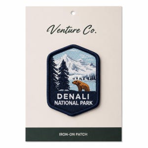 May include: A navy blue iron-on patch featuring a mountain scene with a brown bear and the text "DENALI NATIONAL PARK." The patch is in a dark blue hexagon shape with a light blue border. The words "Venture Co." are written at the top.