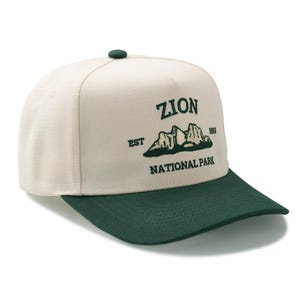 May include: A cream-colored baseball cap with a dark green brim and button. The cap features the text "ZION NATIONAL PARK" in dark green, along with a mountain graphic and the year "1919".