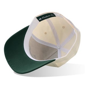 May include: A baseball cap with a cream-colored crown and a dark green brim. The interior features a white sweatband and the text "Venture Co." on a green label.