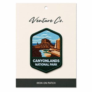 May include: Embroidered Canyonlands National Park iron-on patch. The patch features a landscape scene with a brown mesa, blue sky, and water. The text "CANYONLANDS NATIONAL PARK" is displayed below the image. The patch is on a cream-colored card with the text "Venture Co."