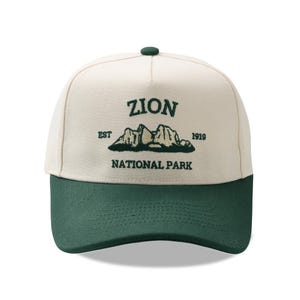 May include: A baseball cap with a cream-colored crown and a dark green brim. The cap features the text "ZION NATIONAL PARK" in dark green, with an illustration of mountains. The text "EST 1919" is also included.