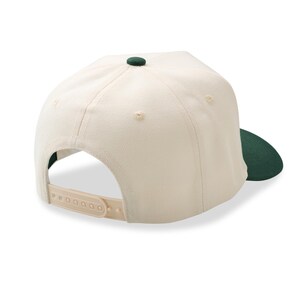 May include: Cream-colored baseball cap with a dark green brim and button. The cap has a snapback closure and a structured design. The back of the cap is visible, showing the adjustable strap.