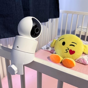 Può includere: Un baby monitor bianco con un obiettivo nero è montato su una sponda di un lettino bianco. Il lettino ha un lenzuolo rosa e un giocattolo di peluche giallo con un viso che fa l'occhiolino e un cuore rosso.