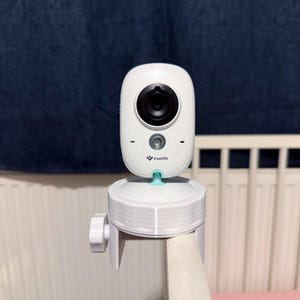 Könnte beinhalten: Eine weiße TrueLife Babyphone-Kamera, die auf einer weißen Kunststoffklemme montiert ist. Die Kameraeinheit ist oval geformt mit einem schwarzen Objektiv, einem kleinen Sensor und dem in Schwarz aufgedruckten Markennamen. Die Klemme hat einen seitlichen Drehknopf.