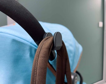 Op de afbeelding: Een close-up van een kinderwagen met een bruine tas die aan een zwarte haak hangt die aan de handgreep is bevestigd. De kinderwagen is lichtblauw en de tas is bruin.