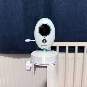 Könnte beinhalten: Ein weiß-hellblaues Babyphone mit schwarzer Kameralinse und kleiner Antenne, montiert auf einer weißen Kunststoff-Klemmhalterung mit Feststellschraube, befestigt an einem weißen Gitterbett. Das Truelife-Logo ist auf dem Gerät aufgedruckt.