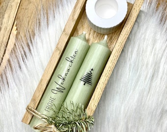 May include: Two green candles with the text "Frohe Weihnachten" and a small Christmas tree design. The candles are in a wooden box with a white ceramic candle holder. A brown tag with the text "Lass es Herzen" is tied to the candles.