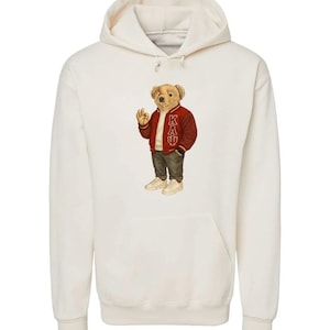 May include: Cream-colored hoodie featuring a bear illustration wearing a red varsity jacket with "KAU" lettering. The bear is also wearing a white shirt, gray pants, and white sneakers. The hoodie has a front pocket and a drawstring hood.