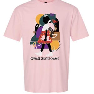 May include: A light pink t-shirt featuring a graphic of a person walking with a colorful abstract background. The person is wearing a white top and a patterned skirt. The text "COURAGE CREATES CHANGE" is printed below the graphic.