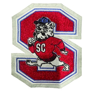 May include: White felt patch with a red felt letter "S" and a white bulldog wearing a red sweater with "SC" on it. The bulldog is wearing a blue baseball cap.