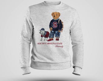 Könnte beinhalten: Hellgraues Sweatshirt mit einem Cartoon-Bären und einer Bulldogge in Outfits. Der Bär hat einen Rucksack und die Bulldogge trägt einen Hut. Der Text "SOUTH CAROLINA STATE University" ist darunter gedruckt.