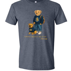 以下が含まれることがあります： ネイビーブルーのTシャツ。クマと犬のイラストが描かれています。クマは青いセーターとジーンズを着ており、犬も同じセーターを着ています。「NORTH CAROLINA A&T STATE University」の文字が下に印刷されています。