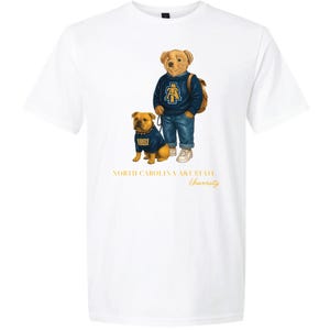 以下が含まれることがあります： お揃いの服を着た漫画のクマとブルドッグが描かれた白いTシャツ。クマは青いセーターとジーンズを着ており、ブルドッグも同じセーターを着ています。「NORTH CAROLINA A&T STATE University」の文字が下に印刷されています。