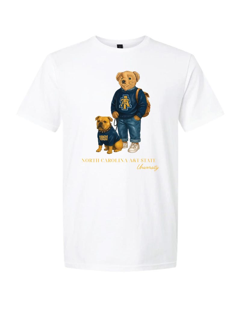 以下が含まれることがあります： お揃いの服を着た漫画のクマとブルドッグが描かれた白いTシャツ。クマは青いセーターとジーンズを着ており、ブルドッグも同じセーターを着ています。「NORTH CAROLINA A&T STATE University」の文字が下に印刷されています。
