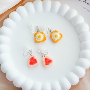 May include: A pair of yellow and orange fried egg earrings and a pair of pink and red strawberry earrings. The earrings are on a white plate with a white background.
