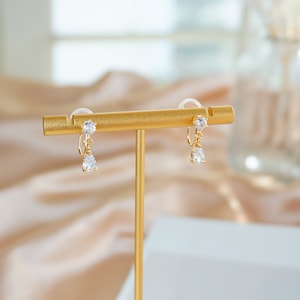 Op de afbeelding: Een paar gouden oorhangers met heldere kristallen. De oorbellen worden gepresenteerd op een gouden standaard.