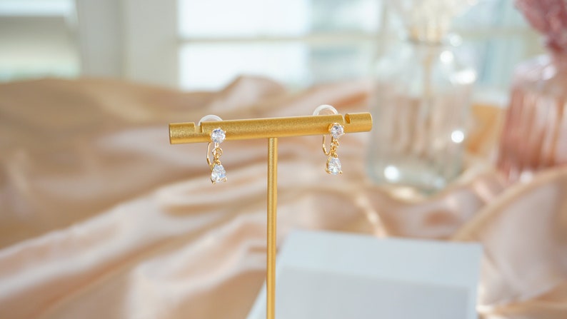 Op de afbeelding: Een paar gouden oorhangers met heldere kristallen. De oorbellen worden gepresenteerd op een gouden standaard.