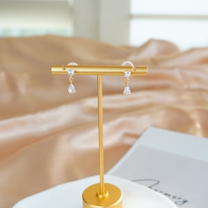 Op de afbeelding: Een paar gouden oorbellen met heldere kristallen druppelaccenten. De oorbellen worden getoond op een gouden standaard tegen een beige satijnen achtergrond.