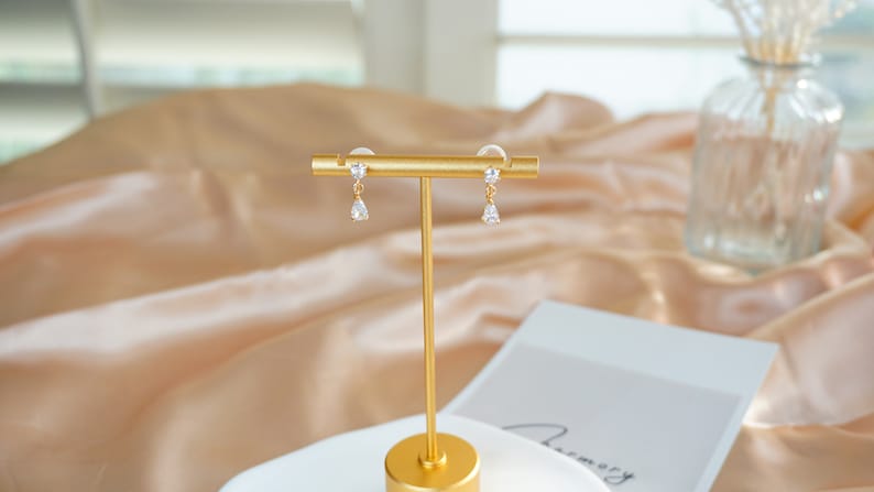 Op de afbeelding: Een paar gouden oorbellen met heldere kristallen druppelaccenten. De oorbellen worden getoond op een gouden standaard tegen een beige satijnen achtergrond.