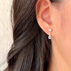 K&ouml;nnte beinhalten: Nahaufnahme eines silbernen Ohrrings mit einem kleinen, runden, klaren Edelstein oben und einem gr&ouml;&szlig;eren, tropfenf&ouml;rmigen, klaren Edelstein, der darunter h&auml;ngt. Der Ohrring wird an einem Ohr getragen, dunkles Haar ist sichtbar.