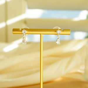 K&ouml;nnte beinhalten: Ein Paar goldfarbene Ohrringe auf einem goldenen Pr&auml;sentationsst&auml;nder. Jeder Ohrring hat einen kleinen, runden, klaren Stein oben und einen tropfenf&ouml;rmigen, klaren Stein, der darunter h&auml;ngt. Der Hintergrund umfasst einen wei&szlig;en, geriffelten Teller und weichen, verschwommenen Stoff.
